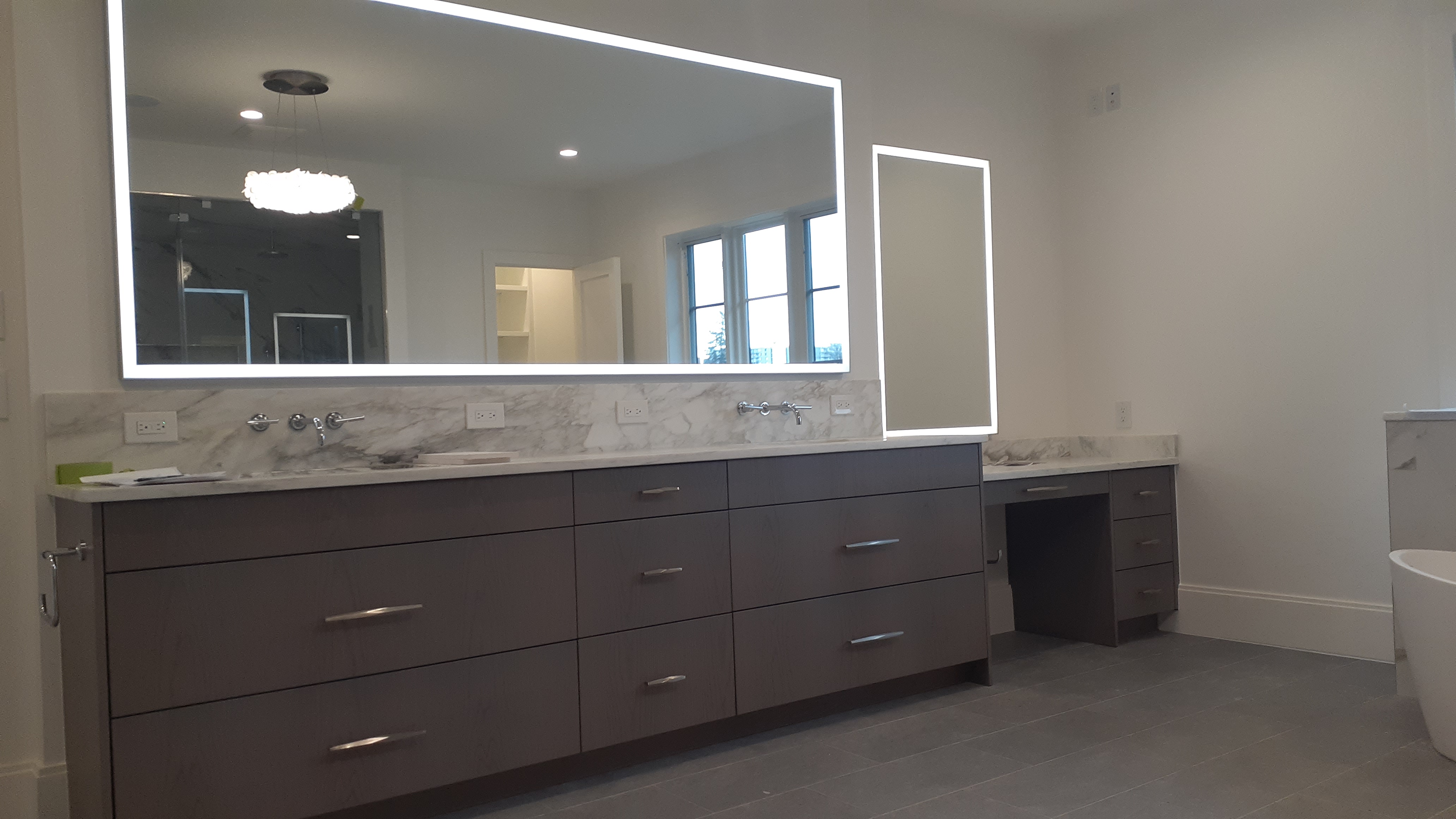 Dark bathroom vanity with flat-panel drawers, marble-look counter, illuminated mirrors, wall-mounted faucets, and a seated makeup station